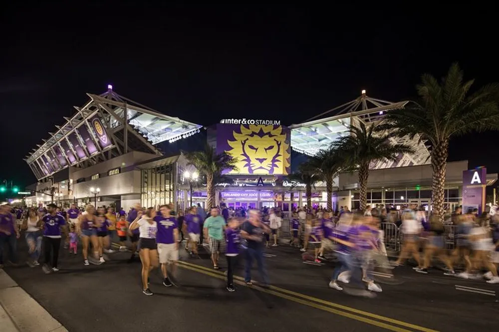 A bustling crowd of people many in purple attire are gathered outside a stadium at night under a sign featuring a lion emblem and the words Orlando City Soccer Club