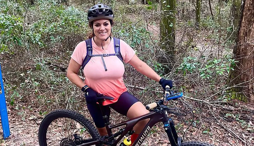 A person is posing with a smile on a mountain bike in a wooded area equipped with a helmet and hydration pack