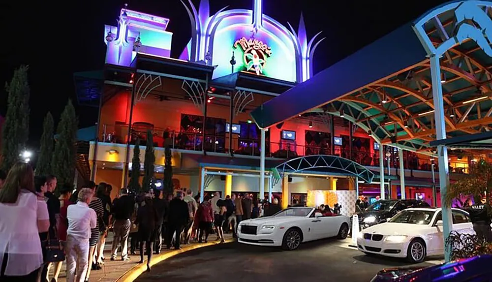 The image shows a bustling nightlife scene with a crowd of people waiting in line outside a brightly lit multi-level entertainment venue and a valet parking service with several cars in the foreground