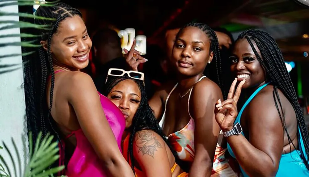 Four women are posing happily for the camera at a social gathering with two of them making peace sign gestures