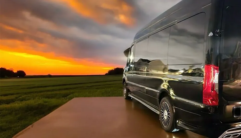 A shiny black van is parked against a vivid sunset sky with a backdrop of green fields