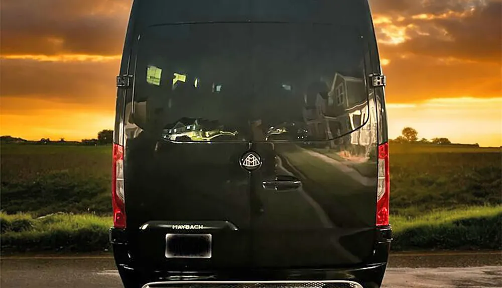 A black van with a reflective surface is parked with a backdrop of a vivid sunset in a rural setting creating a stark contrast between the golden evening sky and the darkened detailed reflection on its rear