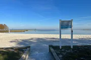 The image shows a tranquil lakeside scene with a sandy shore, a volleyball net, and a sign that likely contains information about the area.