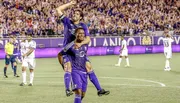 Two soccer players are joyfully celebrating in front of a packed stadium crowd, with one player riding on the other's back.