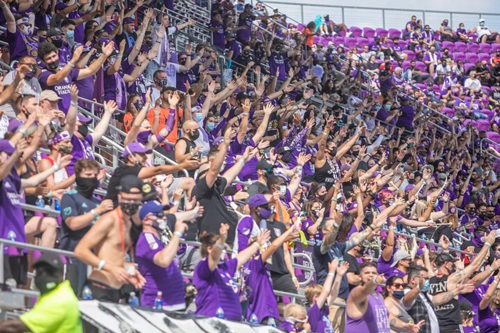 The image shows a lively crowd of sports fans many dressed in purple attire clapping and cheering in the stands of a stadium