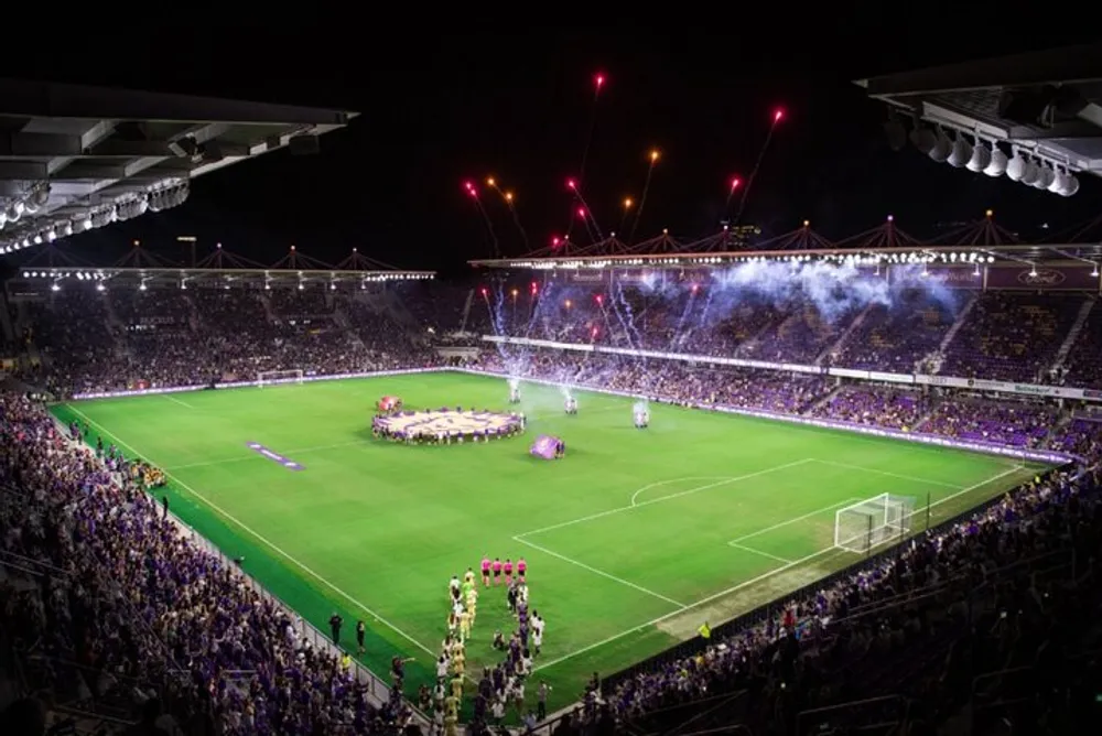 A vibrant football match atmosphere is depicted with a packed stadium purple flares and fireworks lighting up the night