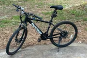 A black mountain bike is parked outdoors on a concrete slab surrounded by grass and leaf litter.