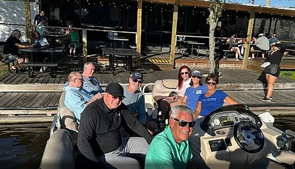 A group of people is smiling for a photo on a boat with a waterfront restaurant in the background