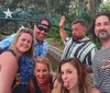 A group of five cheerful adults poses for a lighthearted photo outdoors with a bridge and foliage in the background