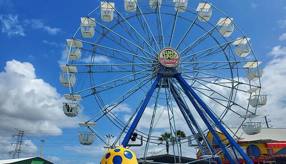 A towering Ferris wheel with white gondolas stands against a backdrop of blue skies and fluffy clouds at an amusement park named Fun Spot America