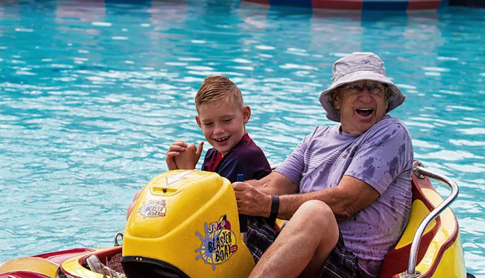 A young boy and an elderly woman are joyfully sharing a ride in a yellow bumper boat on a water attraction