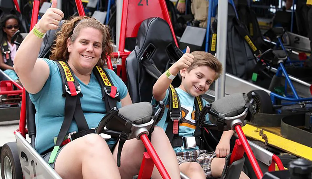 A woman and a young boy are giving thumbs-up signs while seated with safety harnesses in go-karts appearing excited and ready to race