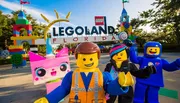 This image features costumed characters resembling LEGO figures in front of the LEGOLAND Florida entrance, with colorful LEGO sculptures and the park's sign in the background.