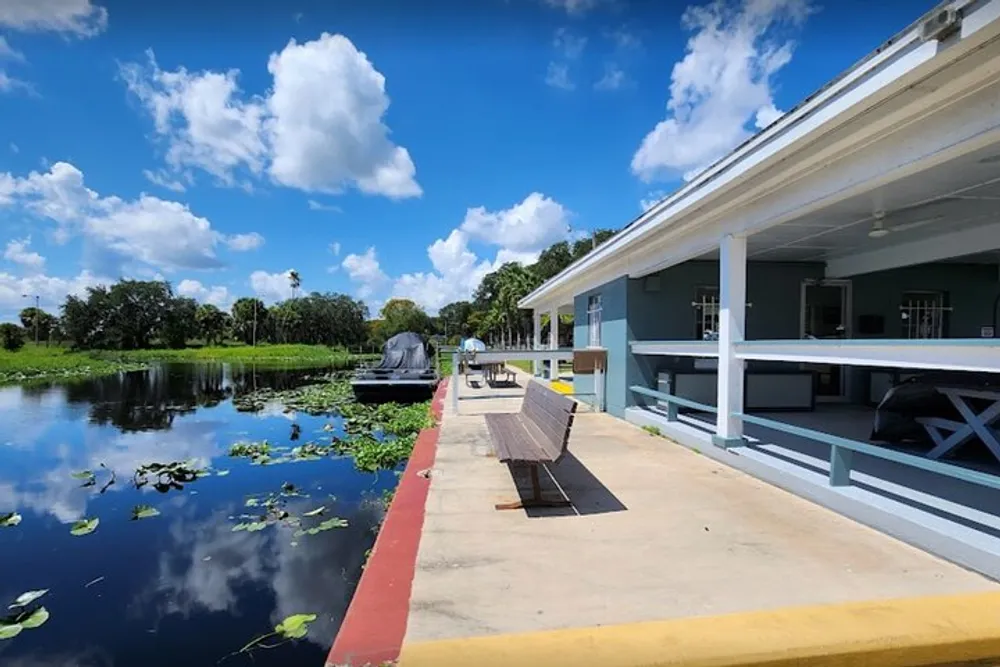 The image shows a serene riverside with an airboat a bench on the dock aquatic plants in the water and a building with a covered porch against a backdrop of a clear blue sky with fluffy clouds