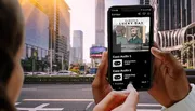 A person is holding a smartphone displaying an interactive story or game with audio options, set against an urban city backdrop.