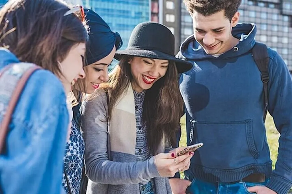 A group of young friends are smiling and looking at a smartphone together outdoors on a sunny day