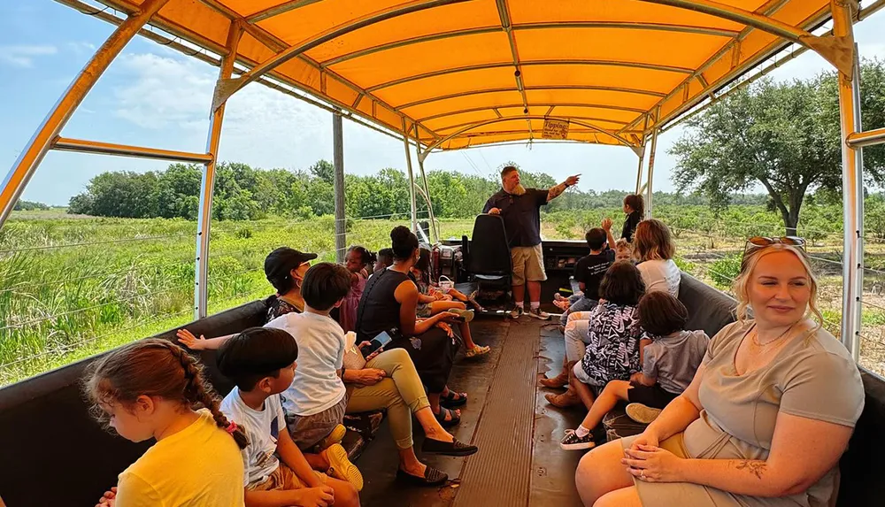 A group of people including children are sitting on a covered open-air vehicle listening to a guide speak and enjoying a scenic view in a grassy area