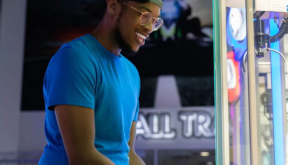 A person wearing a blue shirt and glasses is smiling while playing a claw machine game