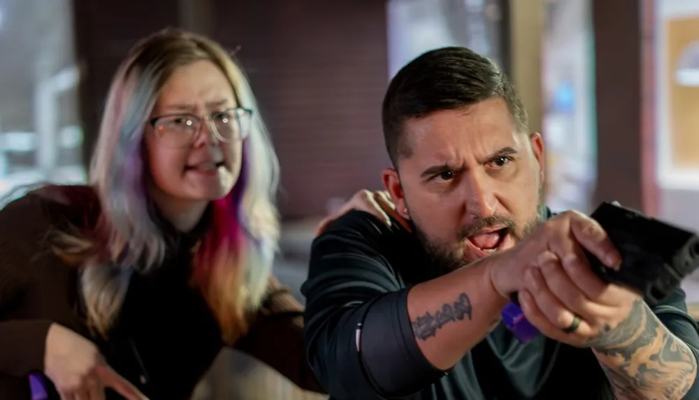 A man with tattoos is aiming a gun while a woman with colorful hair stands beside him both with intense expressions