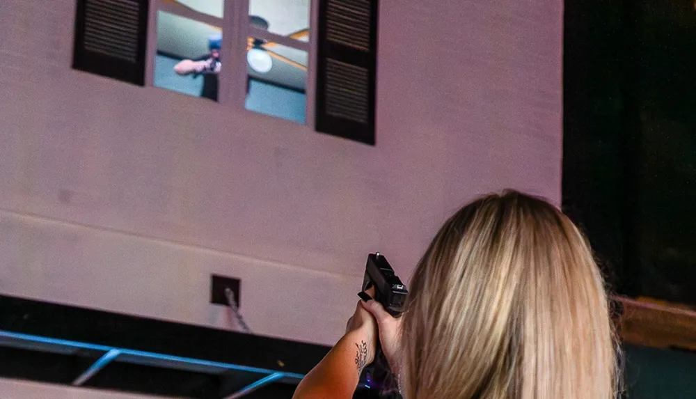 A person standing indoors is aiming a gun at someone visible through a window on an upper floor