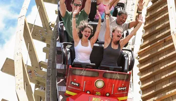 Thrilled passengers with raised arms are experiencing a steep drop on a wooden roller coaster under a clear sky.