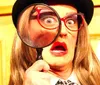 The image shows a person with exaggerated facial expressions wearing a hat and glasses holding a large magnifying glass up to one eye