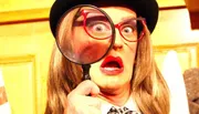 The image shows a person with exaggerated facial expressions, wearing a hat and glasses, holding a large magnifying glass up to one eye.