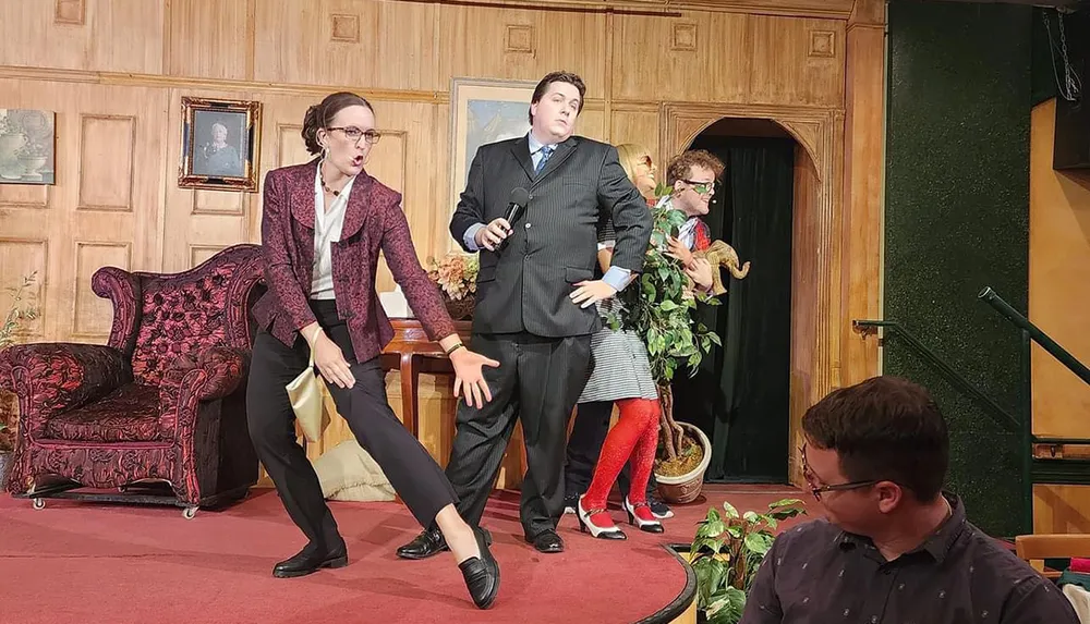 A group of actors appears to be performing a comedic scene on a stage set to resemble a cozy interior complete with furniture and wall paintings