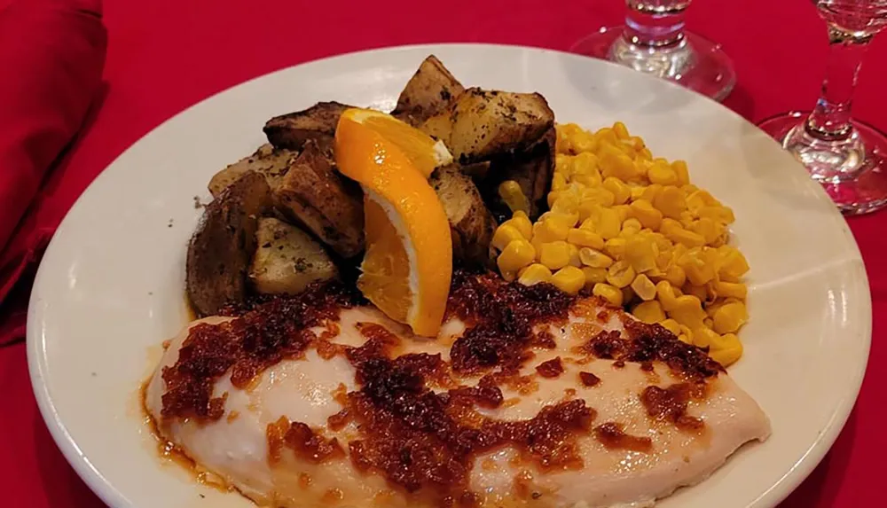 A dinner plate with seasoned roasted potatoes corn kernels and a chicken breast topped with a tomato-based sauce garnished with a slice of orange on a table with a red cloth