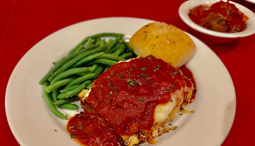 A plate with chicken parmesan green beans and a garlic roll is served alongside a side dish of spaghetti with tomato sauce