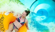A person is smiling and enjoying a ride on a yellow inner tube down a vibrant blue water slide.