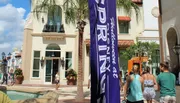 The image captures a sunny day at an outdoor shopping area with visitors walking past a banner, palm trees, and a small fountain.