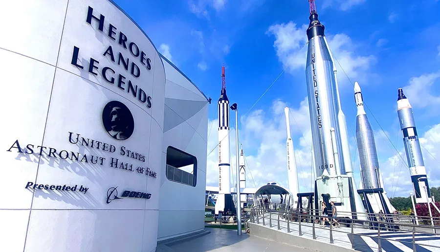 The image features the entrance to the Heroes and Legends United States Astronaut Hall of Fame with several rockets displayed outside against a blue sky.