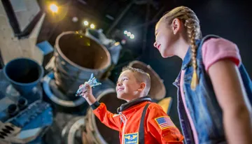 A boy in an astronaut costume holds a toy space shuttle with wide-eyed awe while a girl looks on, both in front of a backdrop of what appears to be a real spacecraft.