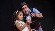 A magician wearing a hat is showing a card trick to a young girl on stage, both looking at the cards she's holding.