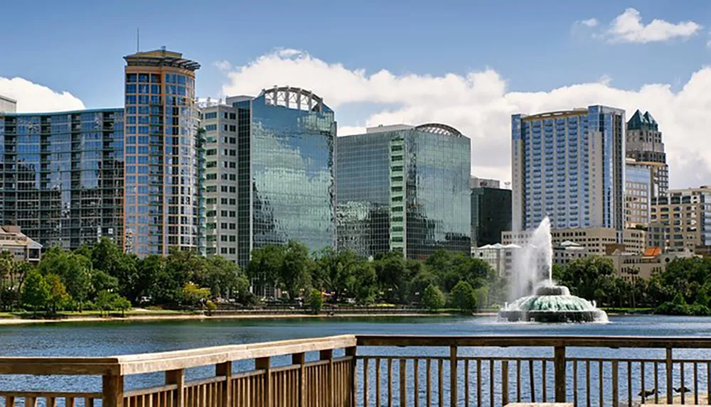 The image shows a tranquil urban park with a lake featuring a fountain in the foreground and a backdrop of modern skyscrapers under a clear sky
