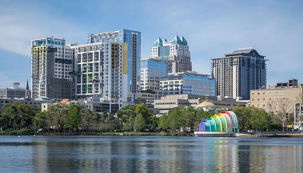 The image shows a modern city skyline across a body of water with a colorful sculptural pavilion near the waters edge