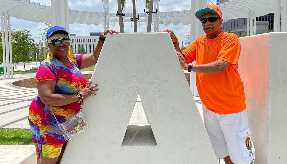 Two people are posing with a large concrete letter A outdoor on a sunny day