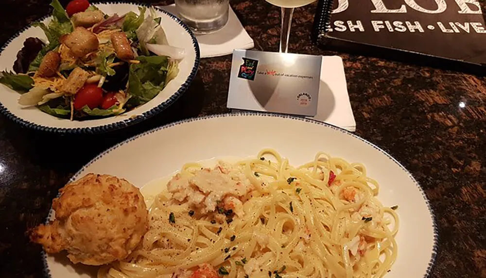 The image shows a plate of spaghetti with a creamy sauce and crab meat alongside a biscuit and a salad with a rack card promoting Eat Play Laugh and a blurred restaurant logo in the background