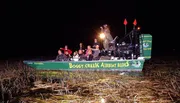 A group of passengers wearing life vests are enjoying a night-time airboat ride through a marshy area with a guide spotlighting the surroundings.