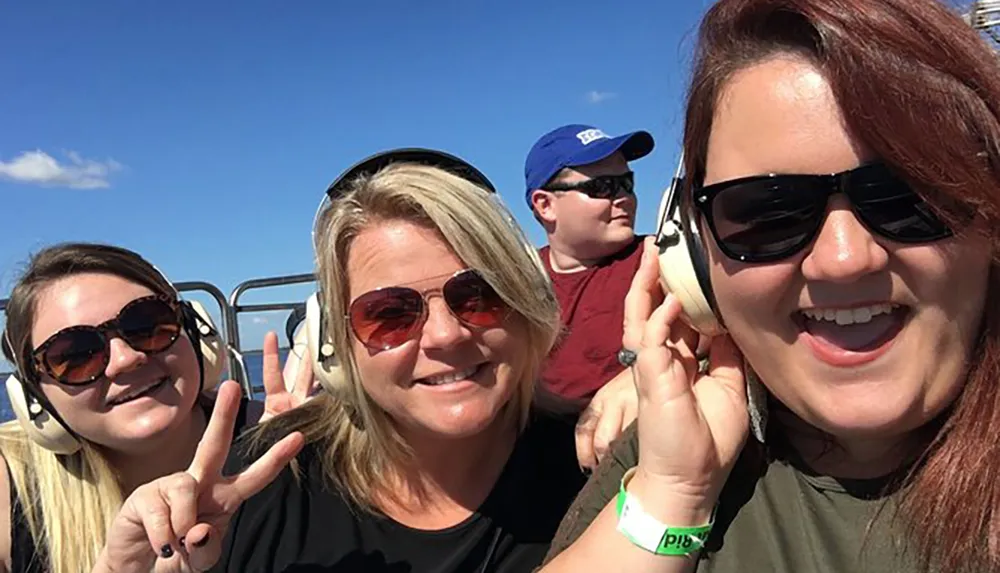 Three people are smiling and taking a selfie while wearing headphones possibly on a tour or an open vehicle under a clear blue sky