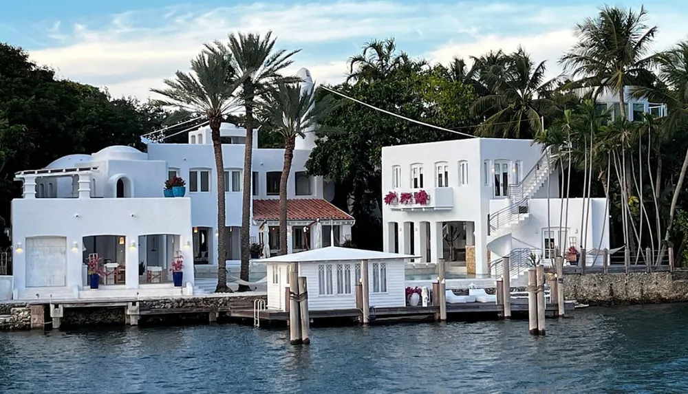The image shows a luxurious white waterfront residence flanked by palm trees with outdoor seating and a private dock