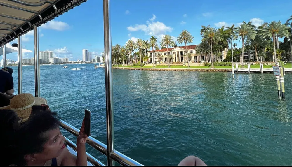 A person takes a photo from a boat showcasing a waterfront view with luxurious houses and palm trees against a backdrop of high-rise buildings