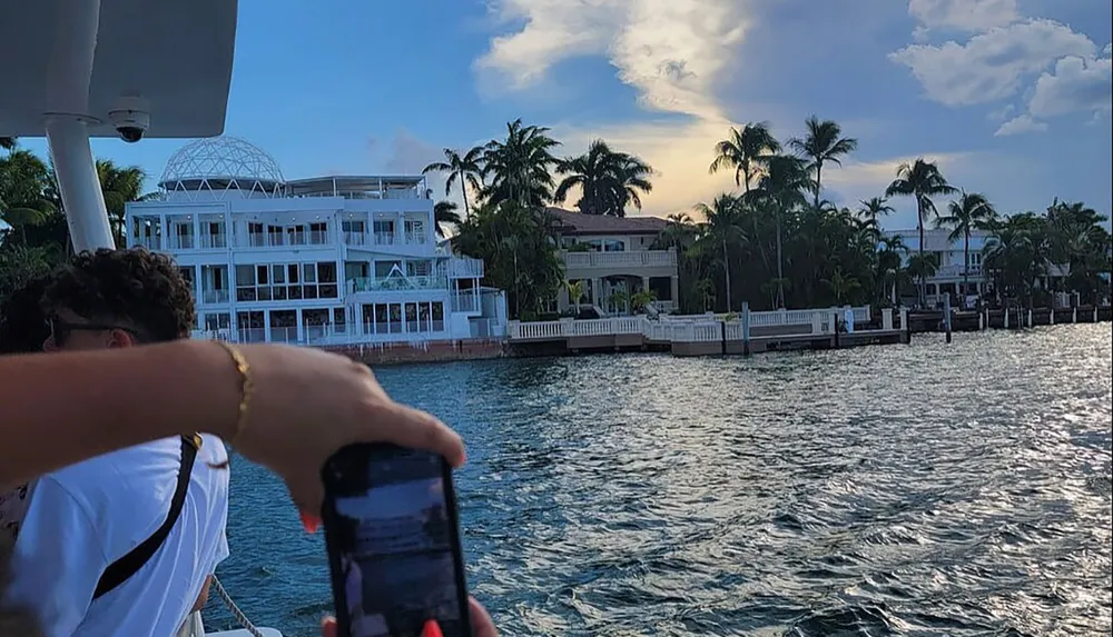 A person is taking a photo with their phone of luxurious waterfront homes under a cloudy sky from a boat