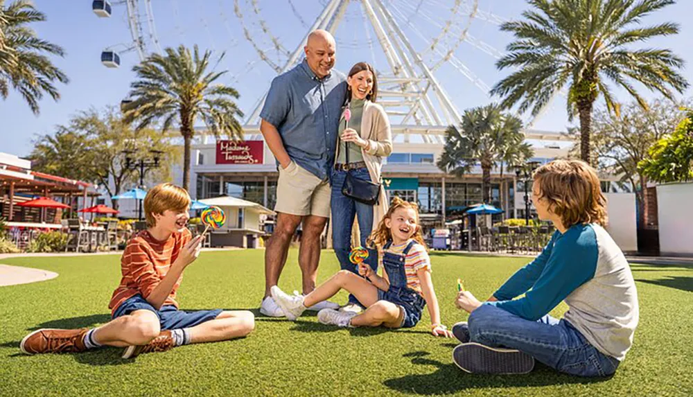 A family appears to be enjoying a sunny day outdoors with two children sitting on the grass holding lollipops and two adults standing behind them all smiling and with a large Ferris wheel in the background