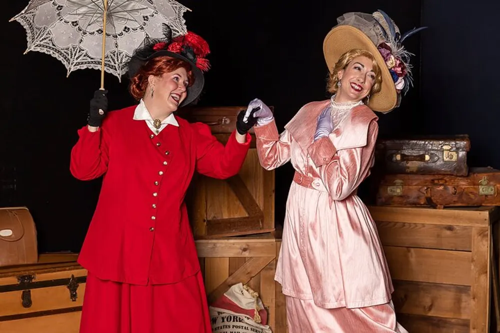 Two women dressed in vintage attire are posing playfully with one holding a lace parasol and the other wearing gloves and a brimmed hat against a backdrop featuring wooden trunks and vintage luggage