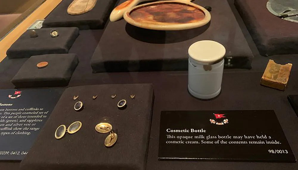 The image shows various antique items on display including jewelry and what appears to be a cosmetic bottle with descriptive labels providing context for museum visitors