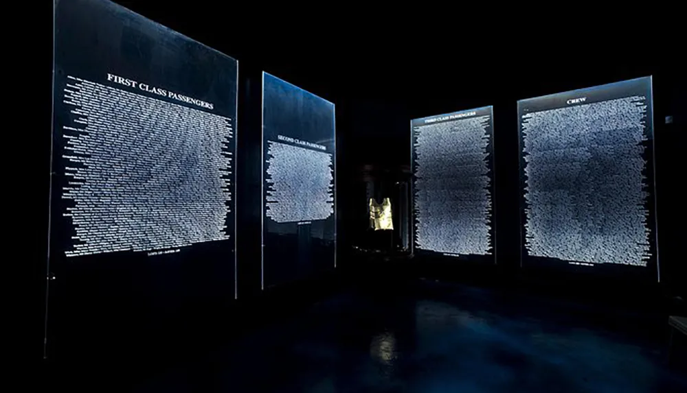 The image displays a dimly lit exhibition with illuminated panels listing the names of first-class passengers second-class passengers third-class passengers and crew likely from a historical voyage or tragic event such as the Titanic disaster