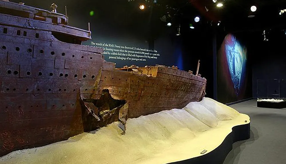 The image shows a museum exhibit featuring a large detailed model of the Titanic shipwreck on a sand-like base with informational text in the background and dim focused lighting creating a somber atmosphere