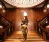 An ornate bronze statue of a figure holding a lamp stands at the base of a grand wooden staircase with detailed craftsmanship
